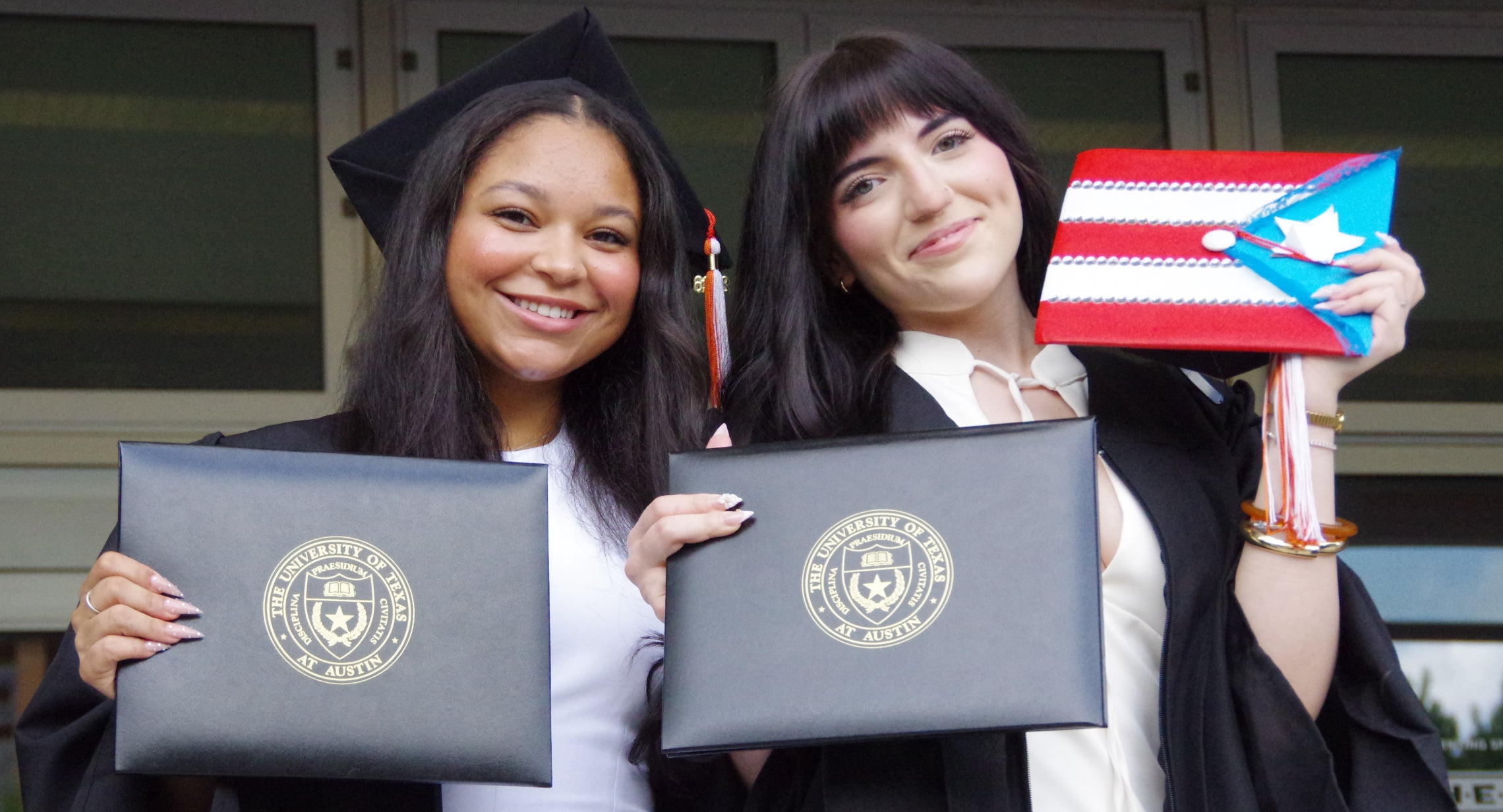 Two graduates in cap and gown proudly holding up their diplomas.