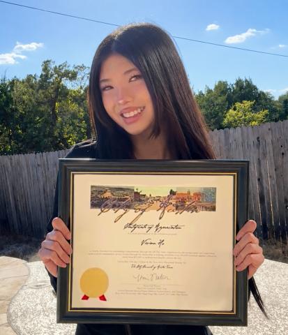Vivian Ho smiles outdoors on a sunny day, holding a framed City of Austin Certificate of Appreciation with a gold seal.