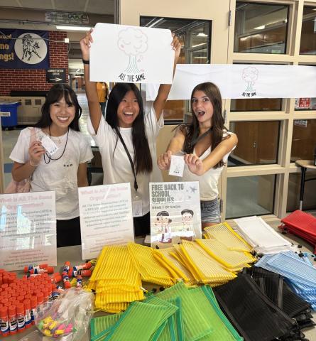 Three smiling teen volunteers in All The Same T-shirts cheer behind a table of free school supplies, with glue sticks and colorful zippered pencil pouches, holding up the organization’s tree logo.