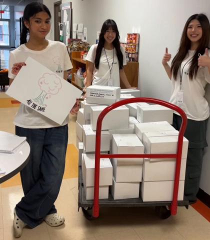Three teen volunteers in All The Same T-shirts stand beside a red cart stacked with white supply boxes in a school hallway, with Vivian Ho on the right giving a thumbs-up.