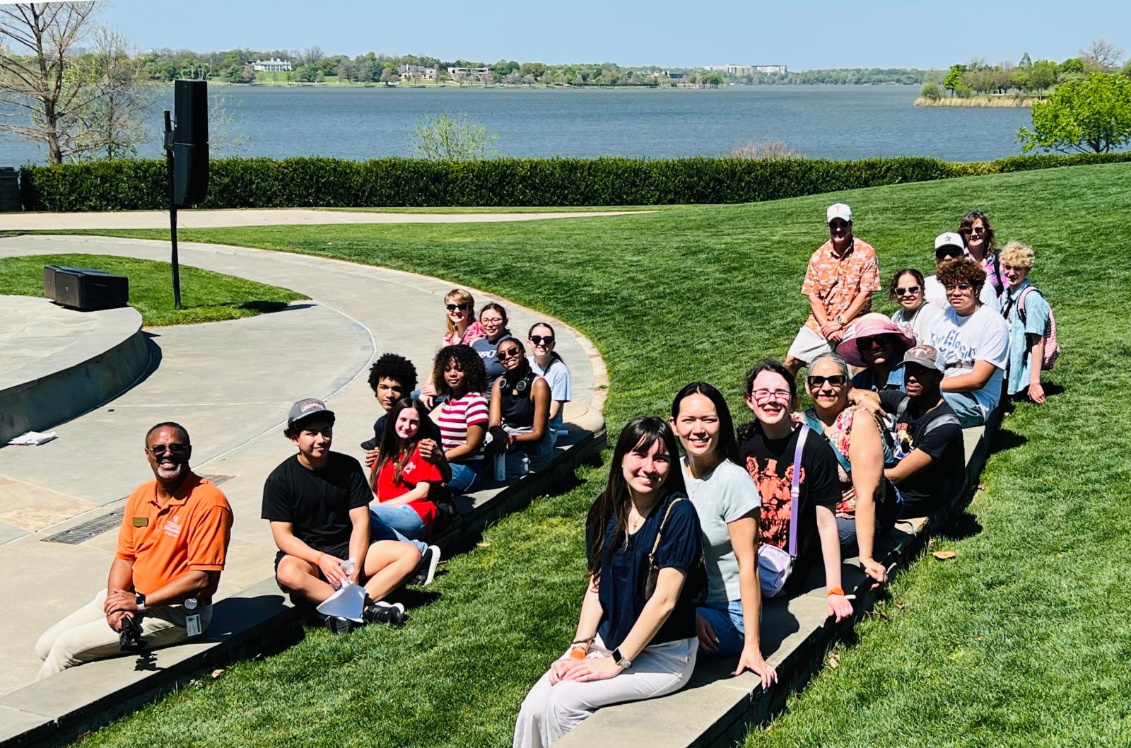Dr. Darnell Horton, in an orange UT polo, sits with about 20 smiling students and adults along the curved stone edge of a lawn, with White Rock Lake behind them.