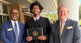 Ian Parham proudly displays his diploma wearing cap and gown, with Principal Darnell Horton and Dan Diehl