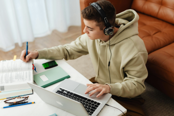 A young man in headphones attending online class at his desk with computer, textbook and notebook