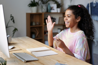 A happy girl sitting at her computer desk waves to her classmates in an online class.