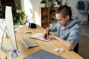 A young man writes in his notebook at his computer desk