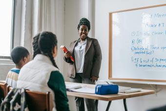 A smiling teacher stands at the whiteboard teaching two students at their desks