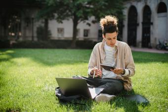 Student studying outdoors on a sunny campus lawn with a laptop and notebook.
