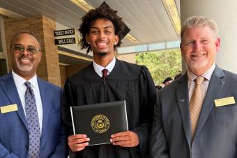Ian Parham proudly displays his diploma wearing cap and gown, with Principal Darnell Horton and Dan Diehl