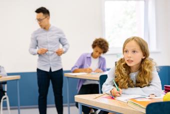 Teacher monitoring a classroom of students writing during a test, with one student looking up from her desk
