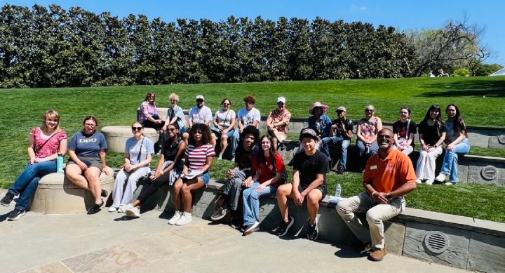 About 20 students and adults sit on tiered stone seating in a garden amphitheater, with Dr. Horton in an orange polo at the front right and tall hedges behind the group.