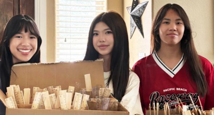 Three teens stand behind open cardboard boxes filled with wooden rulers, with Vivian Ho in the center and co-founder Anika Luangaphay on the left.